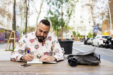 Bearded man sitting outdoors while working over a wooden table - CAVF81981