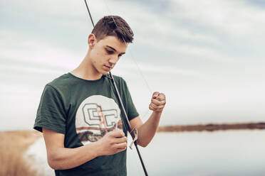 Teenage boy tying hook on fishing rod while standing at lakeshore against  cloudy sky stock photo