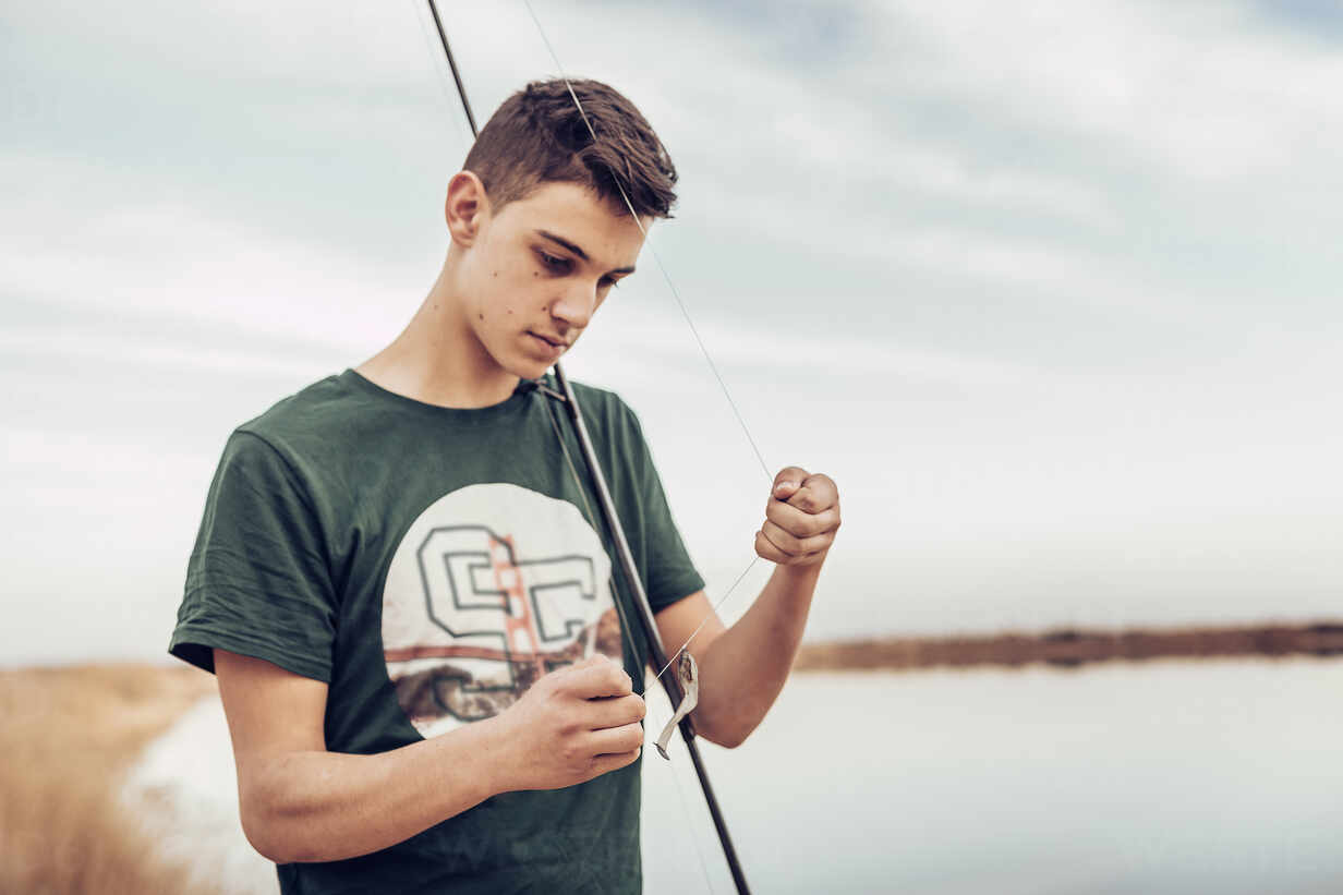 Teenage boy tying hook on fishing rod while standing at lakeshore