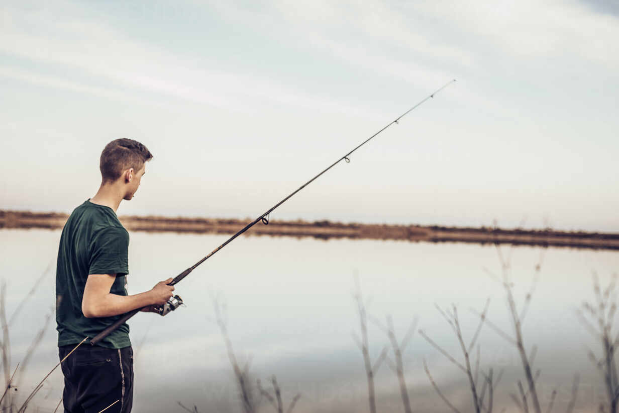 Side view of teenage boy fishing with rod in lake while standing against  cloudy sky stock photo