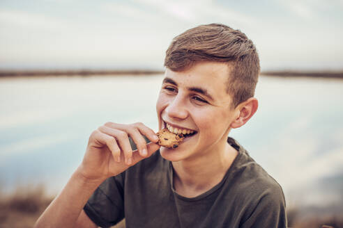 Nahaufnahme eines fröhlichen Teenagers, der einen Keks isst, mit See und Himmel im Hintergrund - ACPF00716