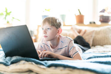 Teenager, der an einem Laptop arbeitet, während er in einem hell erleuchteten Raum auf dem Bett liegt - CAVF82001