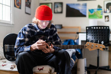 Teenager schaut auf sein Smartphone und hält eine Bassgitarre im Schlafzimmer - CAVF82007
