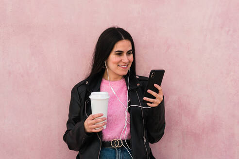 Junge Frau hört Musik und sieht ihr Smartphone in einem rosa Hintergrund - CAVF82016