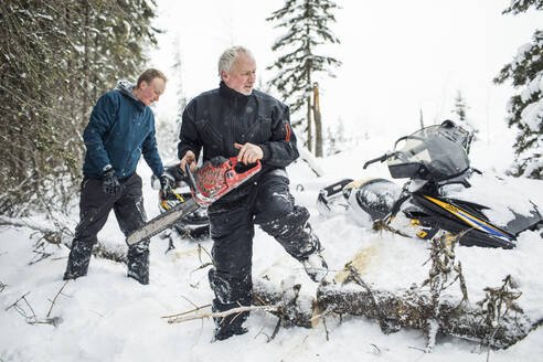 Ein Mann im Ruhestand und sein Sohn benutzen beim Schneemobilfahren eine Kettensäge, um Wege zu räumen. - CAVF82033