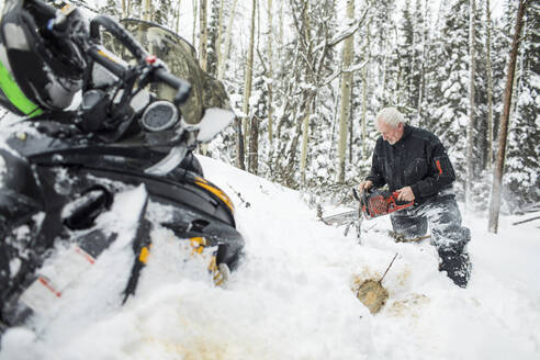 Älterer Mann im Ruhestand, der beim Schneemobilfahren mit der Kettensäge Wege räumt. - CAVF82034