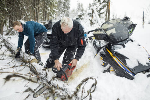 Ein älterer Mann und sein Sohn benutzen eine Kettensäge, um beim Schneemobilfahren die Wege zu räumen. - CAVF82035