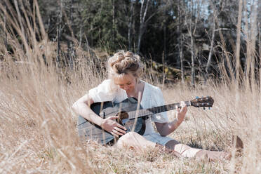 Woman sat in a field happily playing guitar in summer - CAVF82115