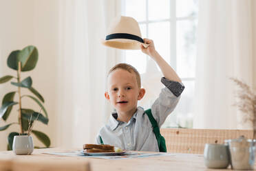Boy holding his hat up whilst eating his lunch at home smiling - CAVF82118