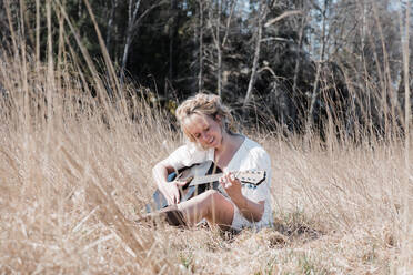 Frau sitzt auf einem Feld und spielt fröhlich Gitarre im Sommer - CAVF82119