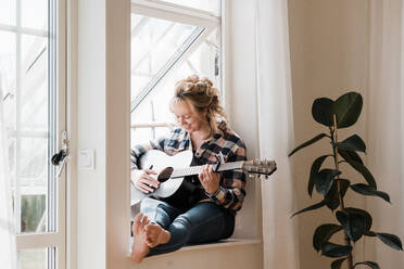 Frau saß zu Hause auf einem Fenstersims und spielte lächelnd Gitarre - CAVF82126