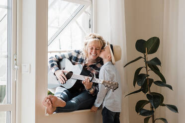 Woman sat playing guitar at home whilst her son gives her a kiss - CAVF82127