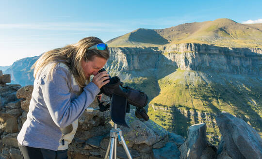 Blonde Frau im Ordesa-Nationalpark, die mit einem Teleskop auf einem Stativ einige Details des Parks betrachtet. Hinter der Frau sind einige Gipfel und Felsen des Panoramas zu sehen. Horizontales Foto - CAVF82134