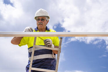 Älterer Techniker, der auf einer Leiter stehend das Dach gegen den bewölkten Himmel ausmisst - DLTSF00693