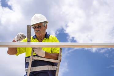 Senior male technician measuring roof while standing on ladder against cloudy sky - DLTSF00695