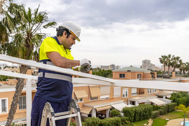 Mature male technician installing solar panel on roof while standing against sky - DLTSF00696
