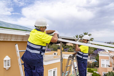 Männliche Mitarbeiter bei der Installation von Solarzellen auf dem Hausdach, während sie über Leitern gegen den bewölkten Himmel stehen - DLTSF00697