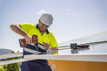 Lächelnder reifer Techniker bei der Installation eines Solarmoduls mit Bohrer auf dem Hausdach gegen den Himmel - DLTSF00701