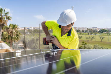 Männlicher Techniker bei der Installation eines Solarmoduls mit einer Bohrmaschine auf dem Hausdach gegen den Himmel an einem sonnigen Tag - DLTSF00705