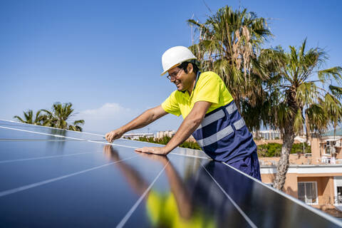 Ein lächelnder, reifer Techniker installiert ein Solarmodul auf einem Hausdach vor blauem Himmel, lizenzfreies Stockfoto