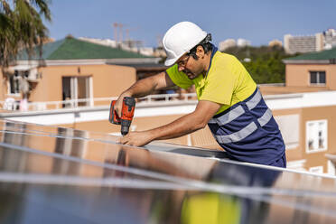 Älterer Techniker bei der Installation eines Solarpanels mit einer Bohrmaschine auf dem Hausdach an einem sonnigen Tag - DLTSF00711