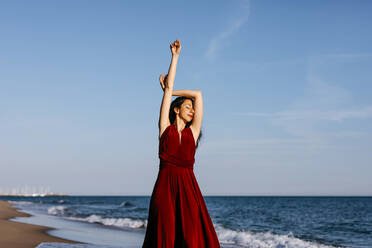 Delicate woman in red dress dancing at the sea, feeling the sun - TCEF00677
