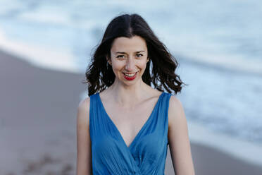 Darkhaired woman in blue dress walking on the beach, smiling, portrait - TCEF00691
