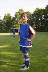 Portrait of confident soccer boy with hands on hips standing on field - AUF00513