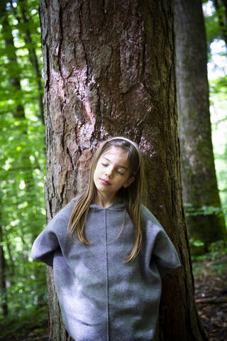 Young girl with eyes closed standing against tree trunk in forest stock photo