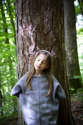 Young girl with eyes closed standing against tree trunk in forest - LVF08904