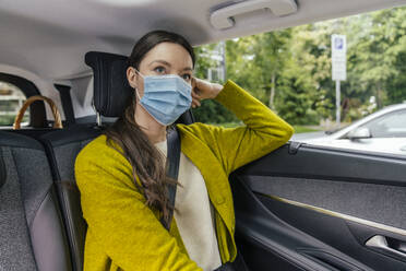 Portrait of pensive young woman wearing protective mask sitting on back seat - MFF05580