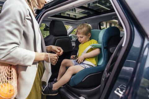 Kleiner Junge beim Befestigen der Sitzglocke des Kindersitzes im Auto, während seine Mutter ihn beobachtet, lizenzfreies Stockfoto