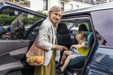 Porträt einer lächelnden Frau, die vor einem Auto wartet, während ihr kleiner Sohn die Sitzglocke des Kindersitzes befestigt - MFF05590