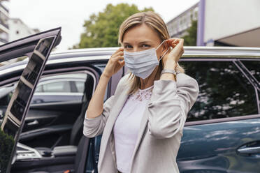 Portrait of woman standing in front of her car putting on protective mask - MFF05603