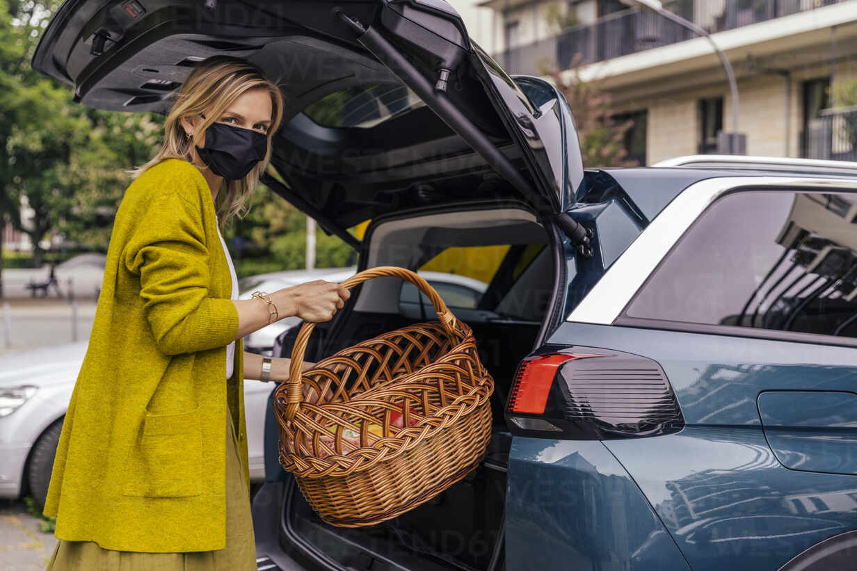 Porträt einer Frau mit Schutzmaske, die ihren Einkaufskorb in den Kofferraum  ihres Autos stellt, lizenzfreies Stockfoto