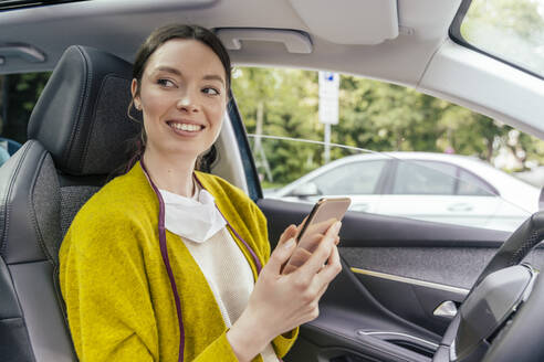 Porträt einer lächelnden Frau im Auto mit Schutzmaske und Smartphone - MFF05615