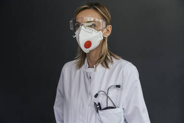 Portrait of doctor wearing FFP3 mask and safety glasses against grey background - MFF05624
