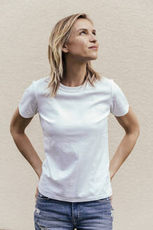 Porträt einer blonden Frau mit weißem T-Shirt, die vor einer hellen Wand steht und nach oben schaut - MFF05634