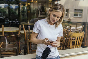 Portrait of woman with protective mask in her hand text messaging in front of a closed coffee shop - MFF05640