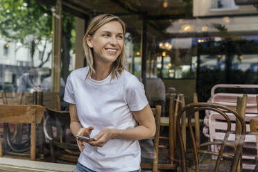 Porträt einer lächelnden Frau mit Smartphone, die vor einem geschlossenen Café steht und in die Ferne schaut - MFF05641