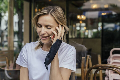 Porträt einer lächelnden Frau am Telefon mit einer Schutzmaske in der Hand vor einem geschlossenen Kaffeehaus, lizenzfreies Stockfoto