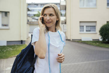 Porträt einer lächelnden blonden Frau am Telefon mit Schutzmaske im Freien - MFF05652