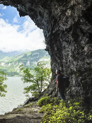 Mature man with backpack standing by rock formation while looking at Lake Como from cave entrance - MCVF00397