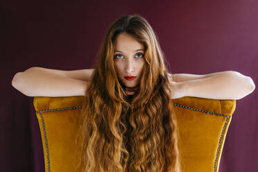 Porträt eines jungen weiblichen Modemodells mit langen braunen gewellten Haaren, das sich auf einen goldenen Stuhl vor einem farbigen Hintergrund stützt - TCEF00703
