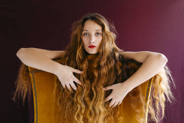 Porträt einer selbstbewussten jungen Frau mit langen braunen gewellten Haaren, die sich auf einen Stuhl vor einem farbigen Hintergrund stützt - TCEF00707