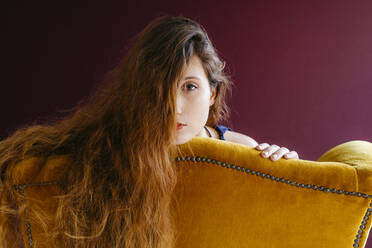 Close-up-Porträt der jungen Frau mit langen braunen Haaren lehnt auf goldenen Stuhl gegen farbigen Hintergrund - TCEF00708