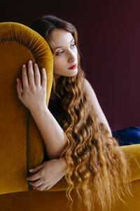 Nachdenkliche junge Frau mit langen braunen gewellten Haaren sitzt auf einem goldenen Stuhl vor einem farbigen Hintergrund - TCEF00709