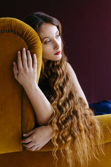 Thoughtful young woman with long brown wavy hair sitting on golden chair against colored background - TCEF00709