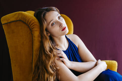 Thoughtful young fashion model with long brown hair looking away while sitting on golden chair against colored background - TCEF00710