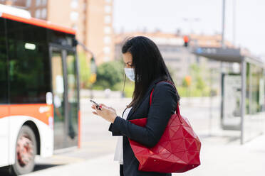 Woman wearing protective mask using smartphone while waiting at bus stop, Spain - DGOF01017